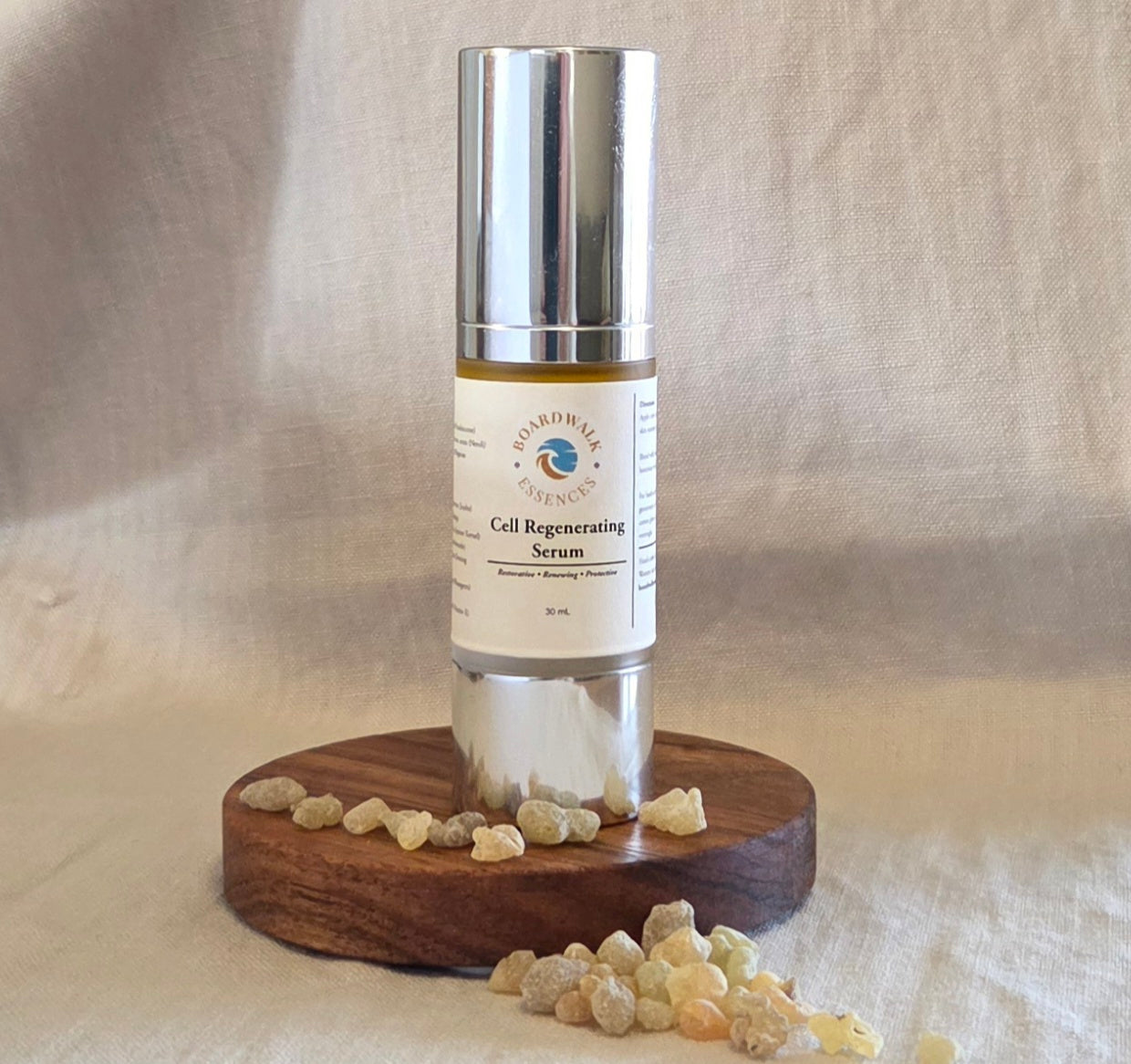  Silver‑capped airless serum bottle with a cream label reading ‘Boardwalk Essences Cell Regenerating Serum – Restorative, Renewing, Protective – 30ml’, placed on a round wooden platform surrounded by small beige resin pieces, against a soft neutral fabric background.
