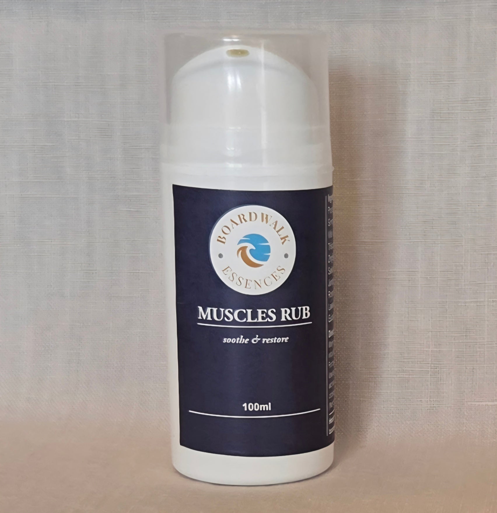   
Amber cylindrical pump bottle with a clear cap, labelled ‘Boardwalk Essences Muscles Rub – soothe & restore – 100ml’, placed on a wooden surface against a soft neutral background.