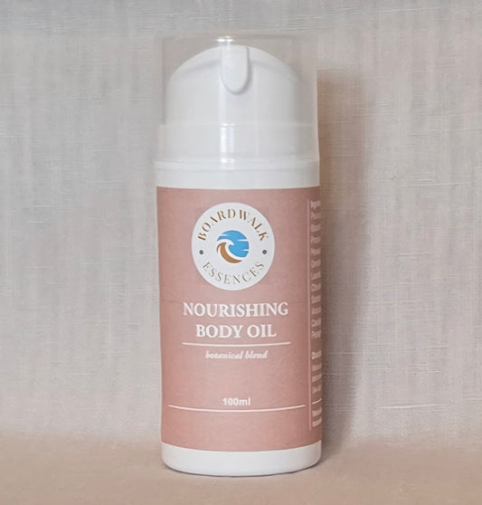 Amber cylindrical pump bottle with a clear cap, labelled ‘Boardwalk Essences Nourishing Body Oil – botanical blend – 100ml’, placed on a wooden surface against a soft neutral background.